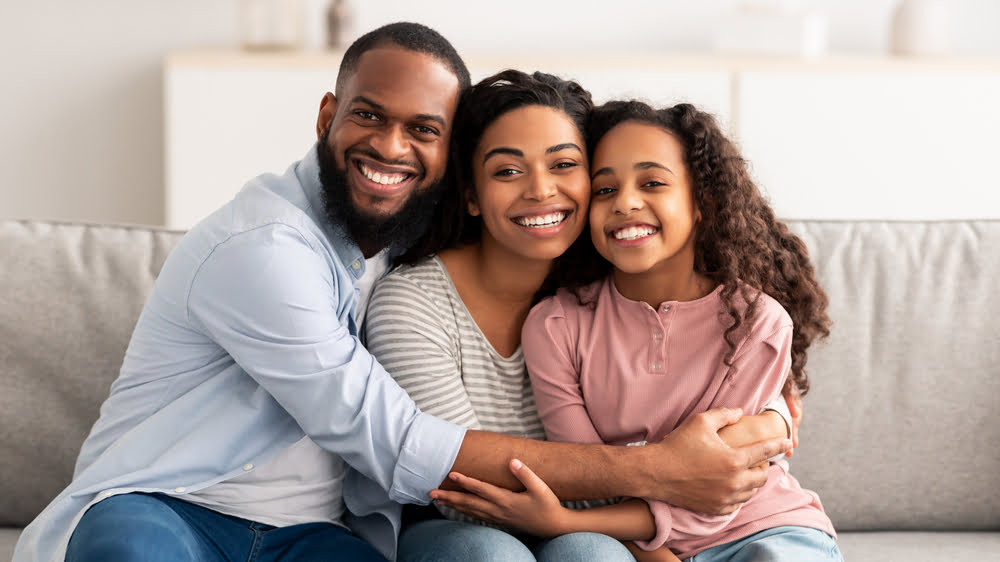 A smiling family of three, sitting on a couch, with the parents hugging their child - Georgina Family Dental A smiling family of three, sitting on a couch, with the parents hugging their child - Georgina Family Dental